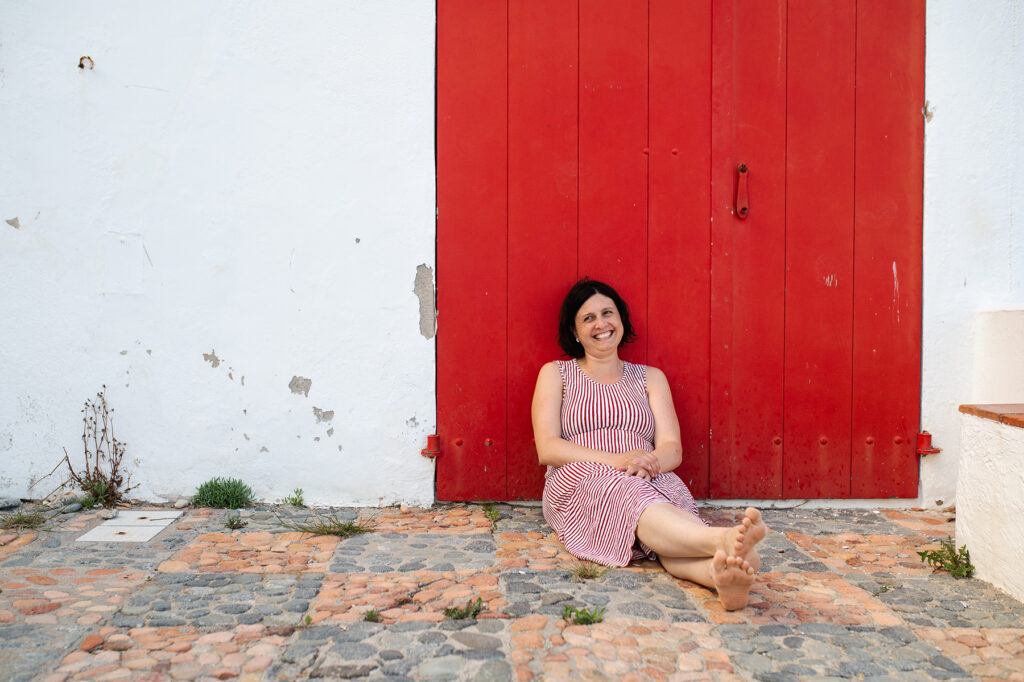 Frau mit rotweißgestreifetem Kleid sitzt vor rotem Tor und lacht bei Fotoshooting